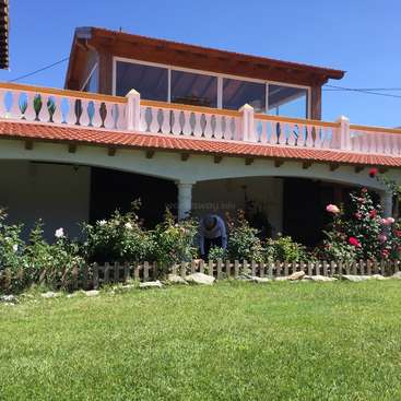 The image depicts a two-story house with a balcony and a garden, featuring a man tending to the plants in the foreground.