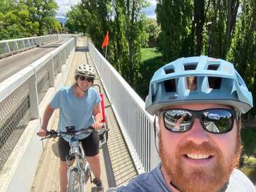 L'image montre un homme et une femme à vélo sur un pont, tous deux portant des casques et des lunettes de soleil, avec des arbres et un drapeau visibles à l'arrière-plan.