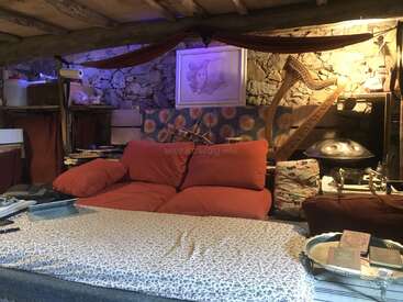 Una acogedora sala rústica con paredes de piedra, sofá rojo, telas estampadas, instrumentos musicales como un arpa y un handpan, estanterías, obras de arte y una cálida iluminación ambiental.