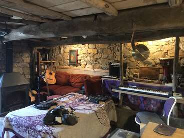 Una acogedora sala rústica con paredes de piedra, vigas de madera vistas, instrumentos musicales, una estufa de leña, un sofá rojo, gatos que duermen y una decoración ecléctica para dar rienda suelta a la creatividad.