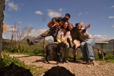 Cuatro amigos y un perro negro posan alegremente al aire libre, uno de ellos con una guitarra en la mano. Detrás de ellos hay montañas, campos verdes, un cielo azul y una gallina.