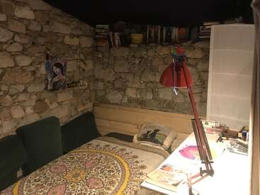 Esta acogedora habitación tiene paredes de piedra, una cama individual con una manta estampada, estanterías, una lámpara roja, un escritorio, un cuadro y varios objetos personales.