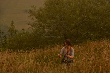 A woman stands in a tall, grassy field surrounded by green trees under a hazy sky, appearing to examine or pick something with her hands.