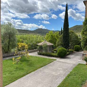 A peaceful garden scene features a small stone structure, lush green lawns, manicured bushes, tall cypress trees, distant hills, and a partly cloudy blue sky.
