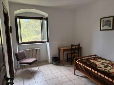This is a simple, tidy room with a single bed, floral blanket, wooden desk and chair, open window, radiator, framed picture, and tiled floor.