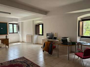 A spacious, light-filled office with tiled flooring, desks, computer, printer, open windows, red chairs, a chalkboard, and a patterned rug on the floor.