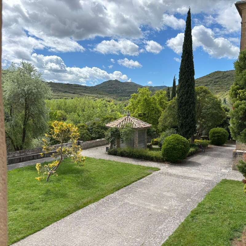A peaceful garden scene features a small stone structure, lush green lawns, manicured bushes, tall cypress trees, distant hills, and a partly cloudy blue sky.