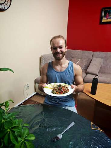 A smiling man in a blue tank top sits indoors, holding a plate of food. There’s a clock, plant, couch, table, and water bottle in the room.