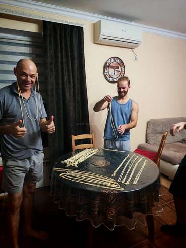 Two smiling men stand in a cozy room, playfully holding long noodles around their necks, with freshly made pasta strands spread out on a round table.
