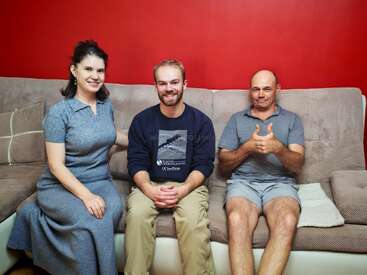 Three people sit on a beige couch with a red wall behind them. They are smiling; the man on the right gives a thumbs up. Cozy, relaxed atmosphere.