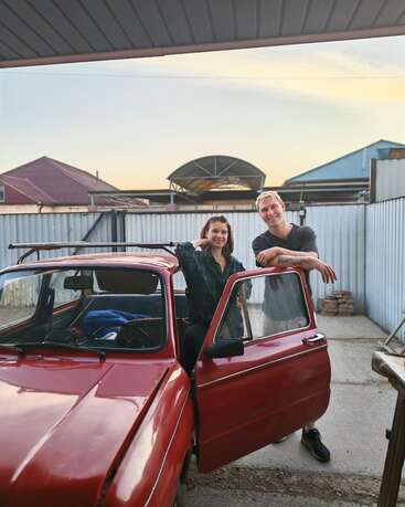 A woman and man stand by a vintage red car in a driveway. The woman is inside, the man outside, both smiling, during sunset. Metal fence surrounds.