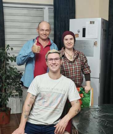 Three people are smiling indoors—two standing, one sitting. The man on the left gives a thumbs-up. There’s a refrigerator and plant in the background. Cozy atmosphere.