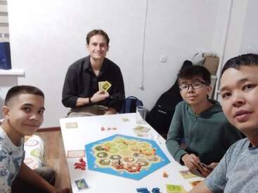 Four friends are playing a board game, Settlers of Catan, around a white table in a cozy, light-colored room. They appear happy and engaged.