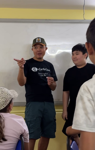 A man in a cap, standing next to a boy, gestures while speaking to a classroom of attentive children, all facing a whiteboard in the background.