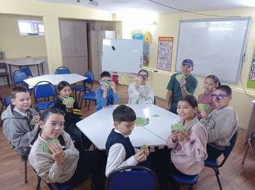 Nine children are sitting around tables in a classroom, playing a card game together, smiling and showing their cards. The atmosphere appears cheerful and engaging.