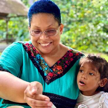Das Bild zeigt eine Frau mit kurzen blauen Haaren und einer Brille, die ein blaugrünes Oberteil trägt und mit einem Kind auf dem Schoß in einem Außenbereich sitzt.