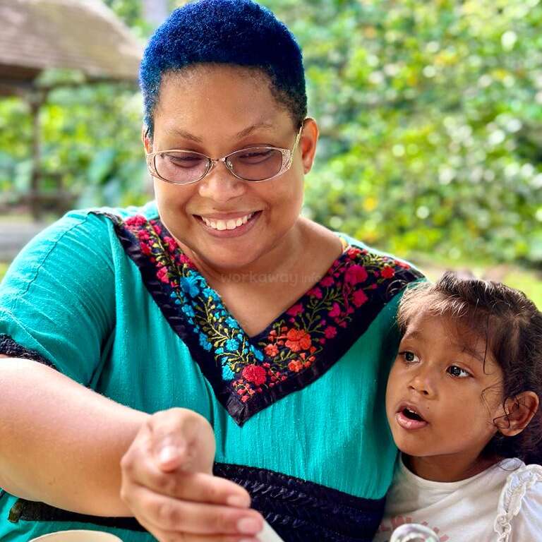 Das Bild zeigt eine Frau mit kurzen blauen Haaren und einer Brille, die ein blaugrünes Oberteil trägt und mit einem Kind auf dem Schoß in einem Außenbereich sitzt.