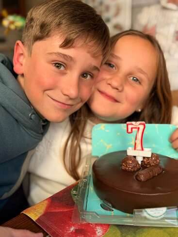 Two smiling children pose closely together with a chocolate birthday cake featuring a number seven candle and chocolates on top, celebrating a special birthday moment.