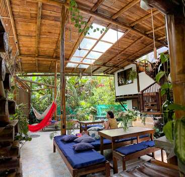 L'image représente un espace intérieur tropical et serein, avec une femme assise à une table, entourée d'une végétation luxuriante et d'un hamac, dégageant un sentiment de détente et de tranquillité.