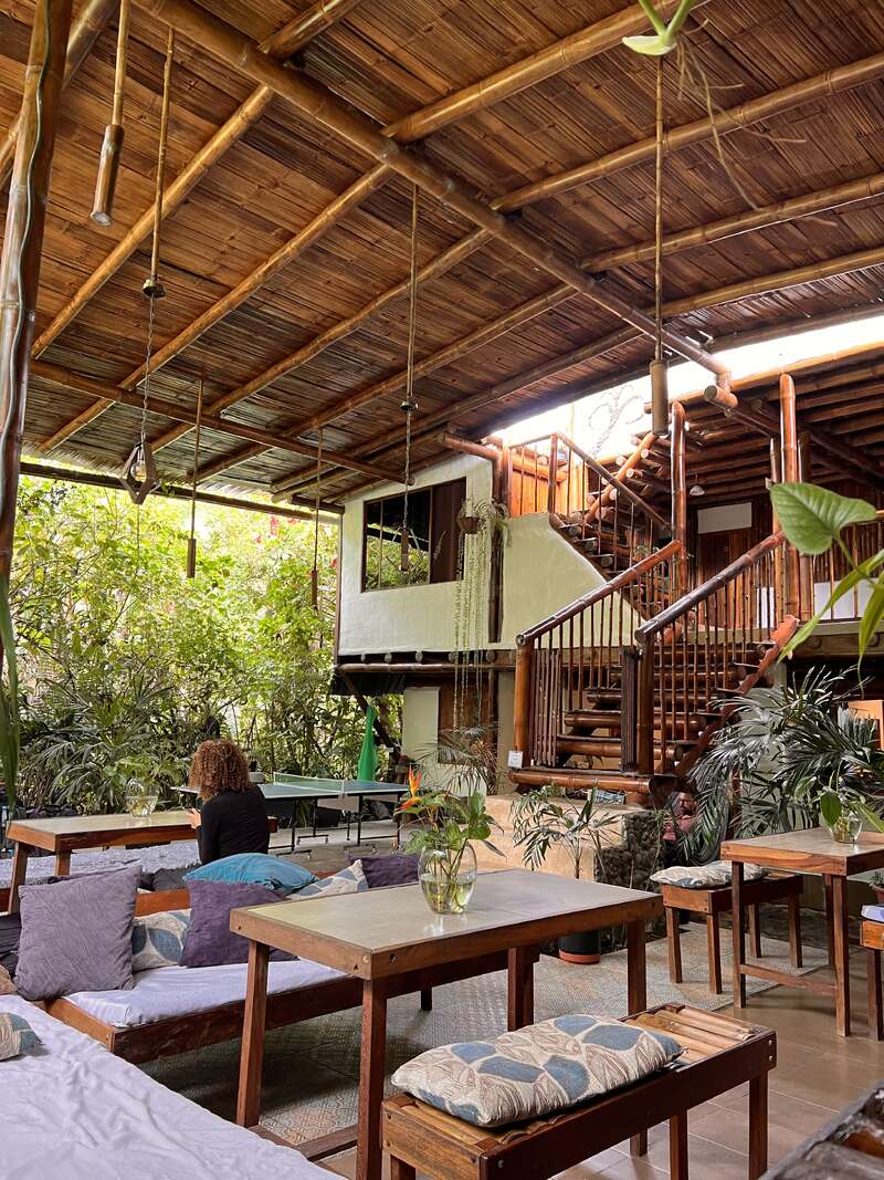 L'image représente un espace intérieur serein avec des tables et des bancs en bois, entouré d'une végétation luxuriante, avec un escalier et une femme assise sur un canapé, dégageant une ambiance naturelle.