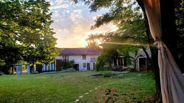 A charming countryside house sits among lush green trees and a spacious yard, bathed in warm golden sunlight at sunset, evoking peace and rustic beauty.