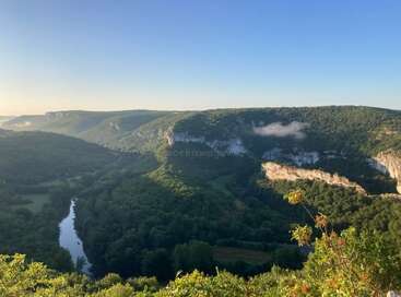 This image shows a peaceful landscape with lush green hills, a winding river, steep cliffs, scattered clouds, and a bright blue sky at sunrise.