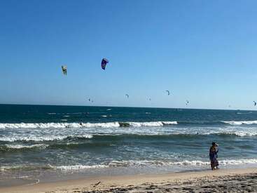 A sunny beach scene with gentle waves, multiple kite surfers in the distance, and a person in a colorful dress standing near the shore, enjoying the view.