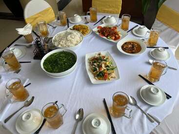 A round table set for six with iced tea, bowls of rice, vegetable dishes, soup, and main courses. White tablecloth and elegant presentation.