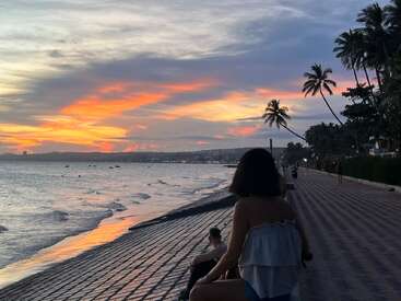 A woman sits on a seaside promenade during a vibrant sunset. Waves gently wash ashore, palm trees line the path, and the sky blazes orange and purple.