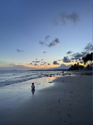 Uma cena tranquila na praia ao pôr do sol, com uma criança brincando na areia molhada perto da beira-mar. Palmeiras e pessoas ao longe se destacam contra o céu colorido.