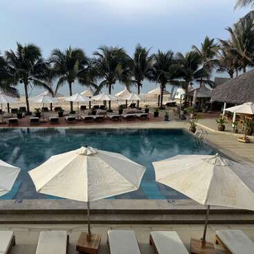 A serene tropical resort features a swimming pool, white sun umbrellas, loungers, palm trees, and an inviting beach with calm waves and soft sunlight in the background.