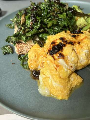 This plate features scrambled eggs topped with a dark sauce, served alongside crispy kale and a slice of seeded bread with a creamy spread underneath.