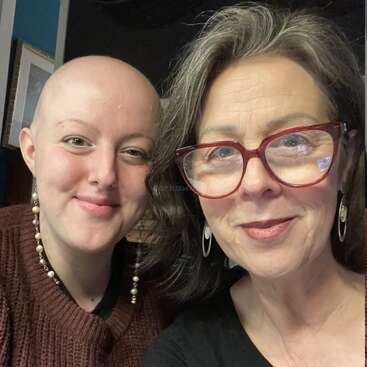 Two women smile warmly for a selfie, one with short gray hair and glasses, the other bald, both wearing earrings and expressing joy, comfort, and connection.