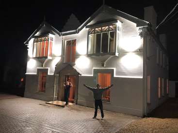 L'image représente deux personnes debout devant une maison blanche, la nuit, l'une faisant un geste vers la maison et l'autre tenant un téléphone.