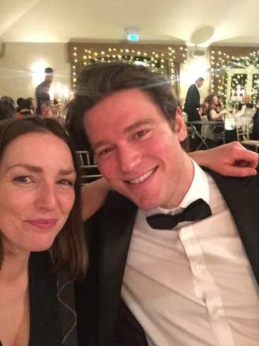 L'image montre un homme et une femme posant pour un selfie lors de ce qui semble être un événement officiel, avec des décorations festives et des personnes en arrière-plan.