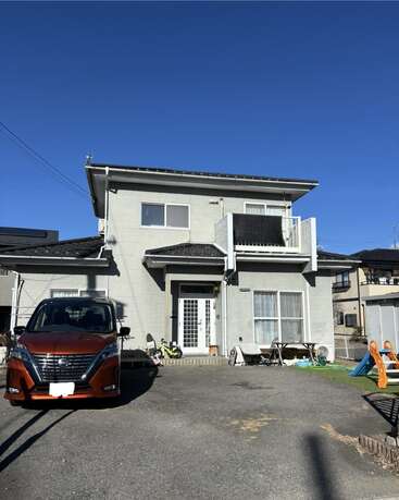 Une maison moderne à deux étages avec un extérieur gris, un balcon et une cour avant. Une voiture orange est garée dans l'allée. Des jouets d'enfants sont éparpillés à l'extérieur.
