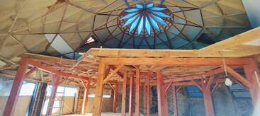 Esta imagen muestra el interior de una cúpula geodésica en construcción, con vigas de madera a la vista, paredes sin terminar, una claraboya en la parte superior y luz natural entrando por ella.