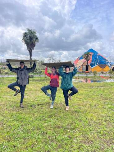 Three people stand on grass, each balancing a board on their heads and posing on one leg like trees, with a geodesic dome and palm tree behind.