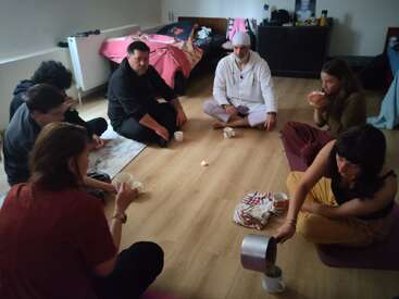 Seven people sit in a circle on a wooden floor, sharing a hot drink together. The setting appears cozy and informal, with beds and casual clothing.
