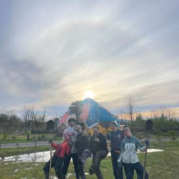 Six people pose joyfully outdoors with shovels in hand. A colorful structure and palm tree stand behind them, with a beautiful cloudy sky at sunset.