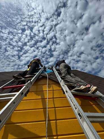 Duas pessoas sobem por escadas encostadas a uma casa amarela, com um céu nublado e dramático acima delas. A perspectiva é de baixo para cima, destacando a altura e o trabalho em equipe envolvidos.