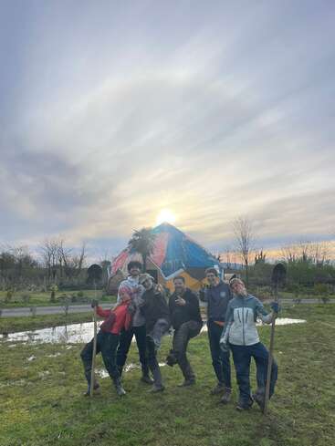 Seis personas posan alegres al aire libre con palas en la mano. Detrás de ellas se ven una estructura de colores y una palmera, con un hermoso cielo nublado al atardecer.