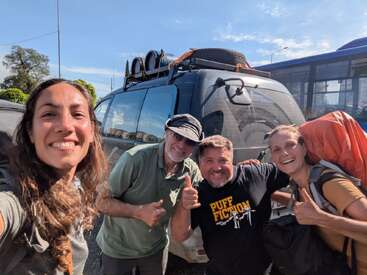 Four smiling people, standing close together, pose for a selfie in front of a van with luggage on the roof. It’s sunny, cheerful, and adventurous.