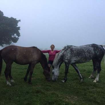 La imagen muestra a una mujer de pie entre dos caballos en un campo cubierto de hierba, con los caballos pastando y la mujer sonriendo a la cámara.
