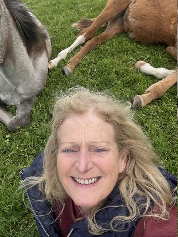A smiling woman with blonde hair takes a selfie on grass, with two horses lying down beside her, creating a joyful and relaxed outdoor atmosphere.