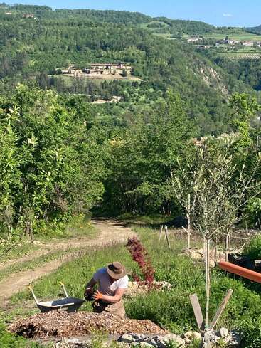 The image depicts a man working in a garden, surrounded by lush greenery and trees, with a picturesque landscape of rolling hills and buildings in the background.