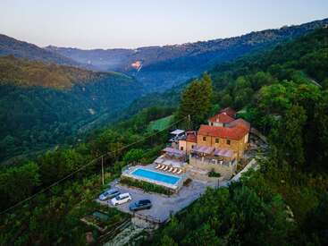 Nestled in lush green hills, a rustic villa with a red roof overlooks a serene pool, parked cars, and offers stunning mountain views at sunset.