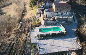 This aerial photo shows a rustic stone house with a red-tiled roof, a rectangular swimming pool, and surrounding trees in a peaceful, rural landscape.
