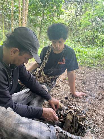 Zwei Menschen sitzen auf dem Waldboden. Der eine schürt ein kleines Lagerfeuer aus Zweigen, während der andere zusieht. Bäume und Grün umgeben sie.