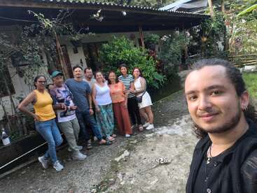 Eine Gruppe von zehn Leuten posiert fröhlich draußen vor einem von Grün umgebenen Haus, wobei eine Person ein Selfie macht und so einen fröhlichen Moment des Beisammenseins festhält.
